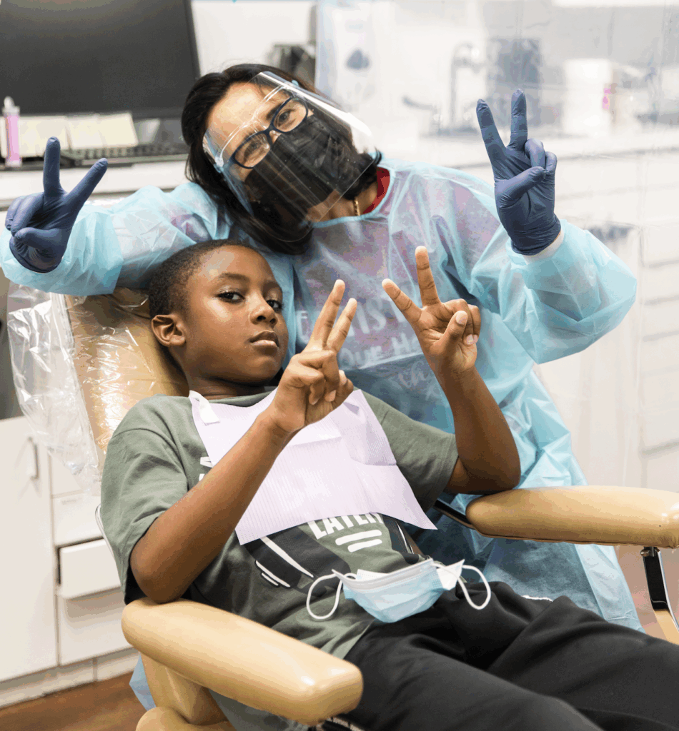 Child in dental chair making peace sign with hygienist in PPE, emphasizing friendly dental care at The Dental Care Group.