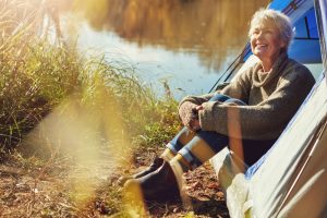 Smiling older woman sitting outside a tent by a river, enjoying nature during a camping trip.