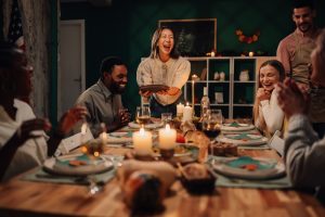 Thanksgiving dinner gathering with a joyful woman serving food at a candlelit table, surrounded by smiling guests enjoying festive dishes, highlighting the importance of holiday meals and dental health.